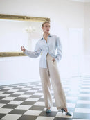 Woman in a light blue shirt and beige pants standing in a room with checkered floor and large mirror.
