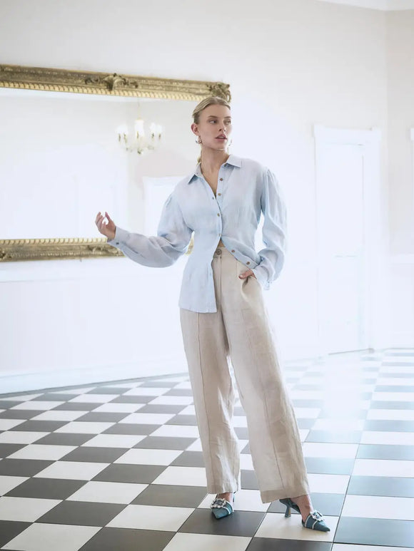 Woman in a light blue shirt and beige pants standing in a room with checkered floor and large mirror.