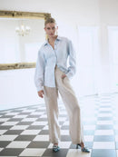 Woman wearing a light blue shirt and beige pants standing in a room with checkered floor and large mirror.