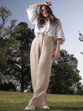 Woman in a white blouse and beige pants standing outdoors with trees in the background