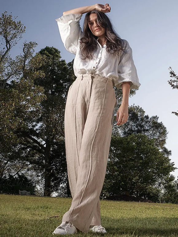 Woman in a white blouse and beige pants standing outdoors with trees in the background